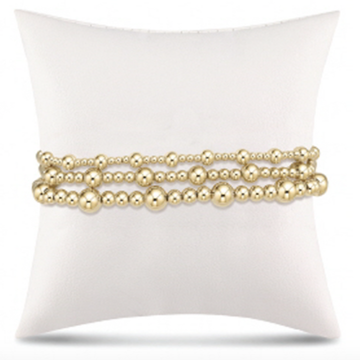 Product photograph of three tiered yellow gold beaded bracelets displayed on a white cushion featuring various sized polished beads and a high-shine reflective finish