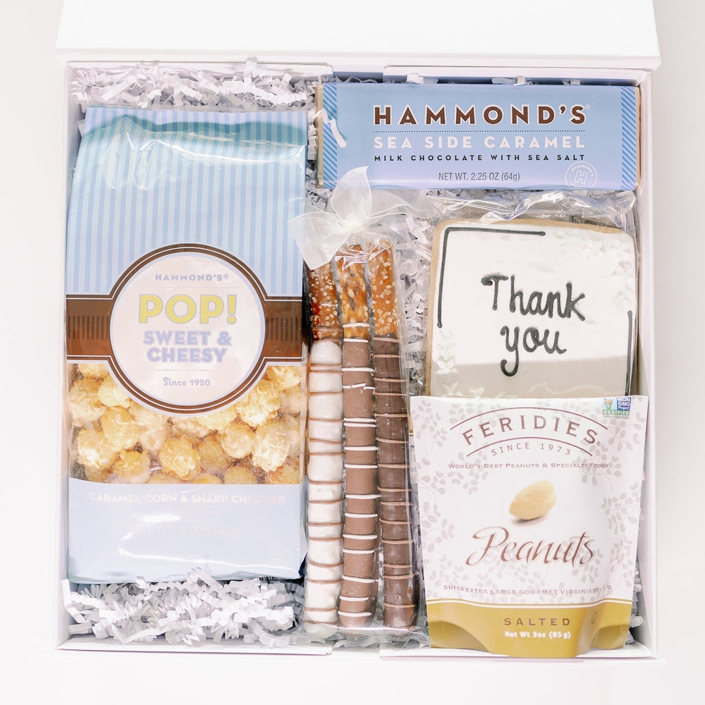Digital photograph of a white gourmet gift box showcasing Hammond's sweet and cheesy popcorn, Feridies salted peanuts, three drizzled chocolate pretzels with a white bow, and a square frosted cookie with Thank You in black script