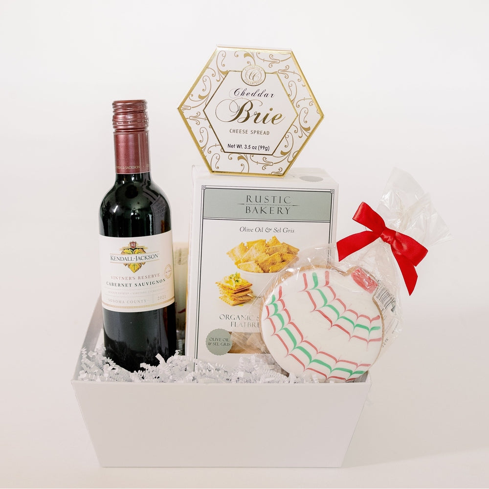 Photograph of a white gourmet gift basket on a studio background featuring a bottle of Kendall-Jackson Cabernet Sauvignon, a gold-trimmed hexagonal cheese spread box, and a large round iced cookie with red and green scalloped patterns tied with a red ribbon
