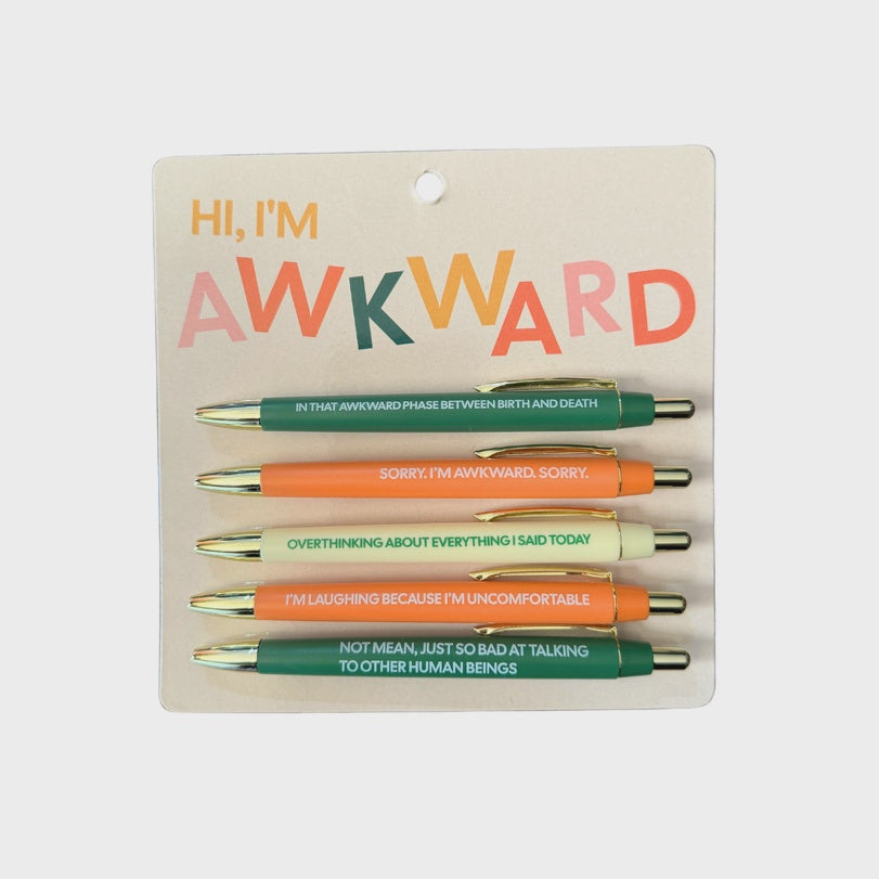 Stationery set by The Found featuring five ballpoint pens with gold hardware and humorous phrases on a tan card with a large colorful HI, I'M AWKWARD header and pen barrels in shades of forest green, orange, and cream