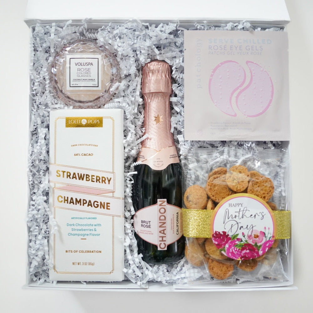 Photograph of a curated Mother's Day gift box featuring a Chandon Brut Rosé bottle, Voluspa candle, and Lolli and Pops chocolate bar nestled in white crinkle paper with a gold glitter ribbon and pink floral sticker