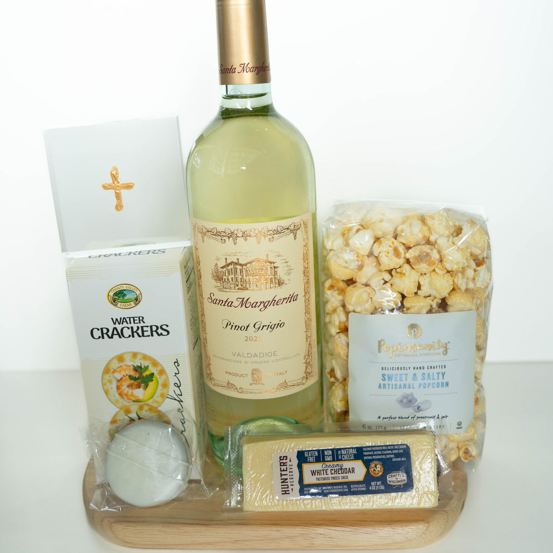 Photograph of a gourmet wine and snack gift set on a wooden board featuring a bottle of 2023 Santa Margherita Pinot Grigio, Hunter's Reserve white cheddar, and Popinsanity popcorn next to a card with a gold foil cross