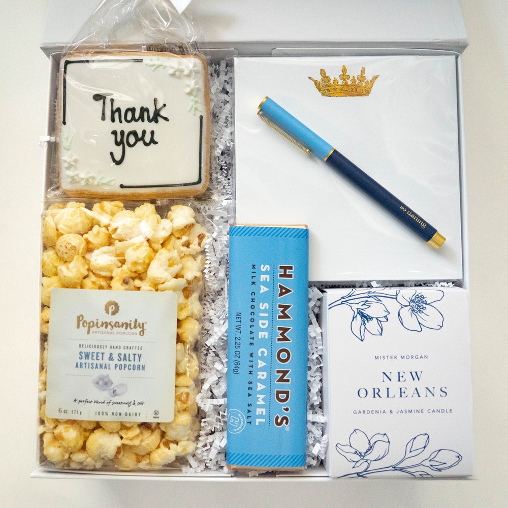Photograph of a curated gift box featuring a hand-lettered Thank You cookie, a bag of Popinsanity popcorn, a blue Hammond's sea salt caramel chocolate bar, and a New Orleans candle box accented by a gold crown stationery set and a navy blue pen