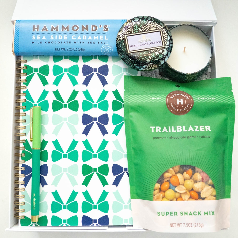 Product photograph of a curated gift box featuring Hammond's and Voluspa products including a vibrant teal and navy bow-patterned notebook, a matching slim green pen, and a decorative floral candle tin set inside a white box
