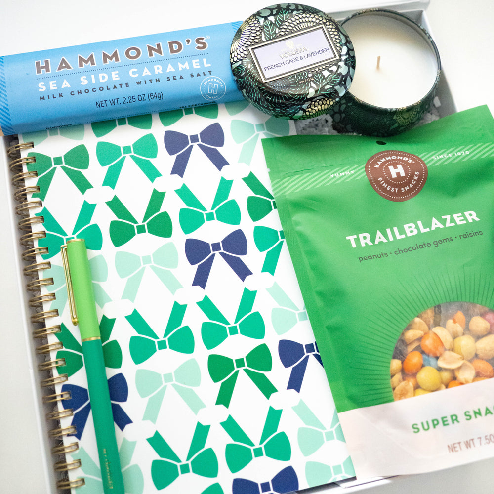 Photograph of a curated gift box containing a Hammond's Sea Side Caramel bar, a Voluspa French Cade and Lavender candle, a Hammond's Trailblazer snack bag, and a notebook featuring a repeating teal and green bow pattern and a matching green pen