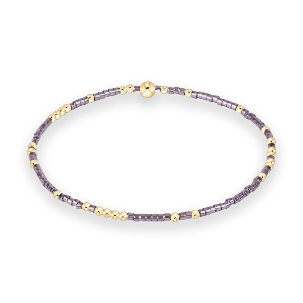 Handcrafted beaded jewelry featuring a circular stretch bracelet on a white background with a large central gold spherical bead, iridescent lavender-grey cylindrical spacers, and polished gold-tone round accents