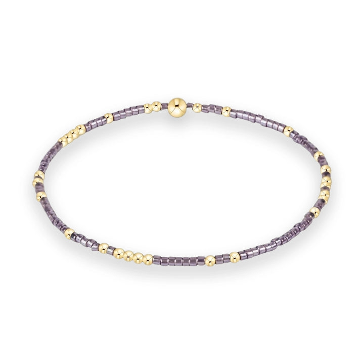 Handcrafted beaded jewelry featuring a circular stretch bracelet on a white background with a large central gold spherical bead, iridescent lavender-grey cylindrical spacers, and polished gold-tone round accents