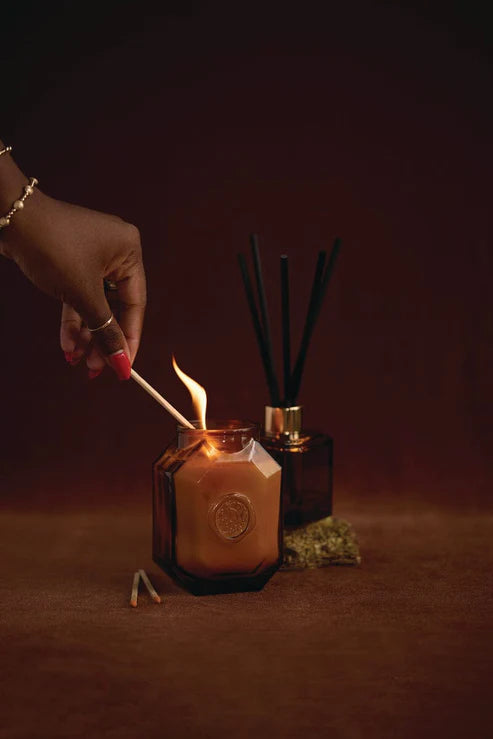Digital photograph of a hand with red manicured nails lighting a match to an amber-colored geometric candle jar in a warm studio setting featuring a flickering orange flame and a black reed diffuser against a dark backdrop