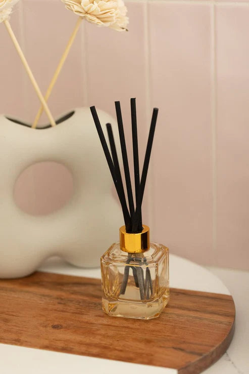 Photograph of a square glass reed diffuser with black reeds and a gold collar on a wooden and marble tray against a pink tiled wall and a cream-colored abstract vase