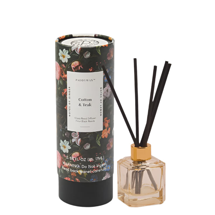 Studio photography by Paddywax showcasing a Cotton & Teak glass reed diffuser set against a white background, featuring a dark cylindrical tube with vintage floral botanical illustrations and a square amber glass bottle with a metallic gold collar and four fanned-out black reeds