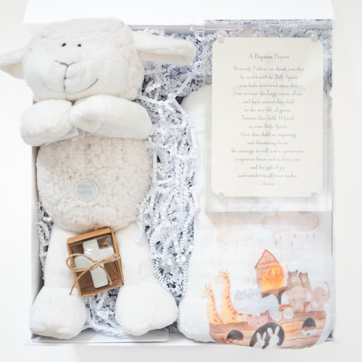 Product photograph of a baptism gift set in a white box featuring a plush white lamb with a Press Here button, a small white cross tied with twine, and a muslin swaddle blanket illustrated with a colorful Noah&