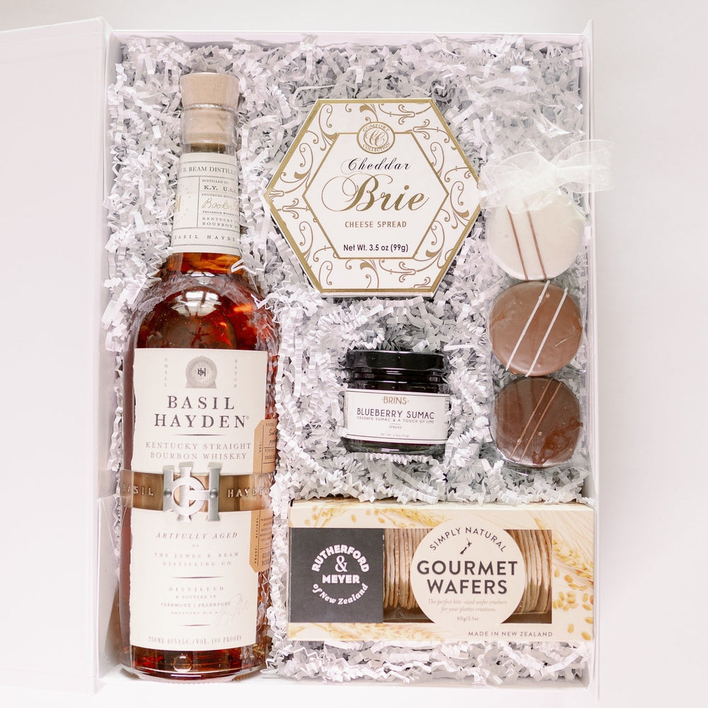 Digital photograph of a curated gourmet gift box featuring a bottle of Basil Hayden Kentucky Straight Bourbon Whiskey, a hexagonal Cheddar Brie cheese spread, and Rutherford & Meyer gourmet wafers nestled in white crinkle paper beside chocolate-covered cookies with white icing stripes