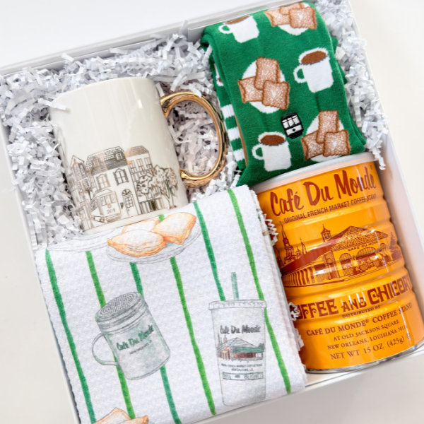 Photograph of a New Orleans gift set featuring the iconic Café Du Monde coffee stand on a bright orange tin, a white ceramic mug with a gold handle and St. Charles streetcar illustration, and green patterned socks decorated with beignets