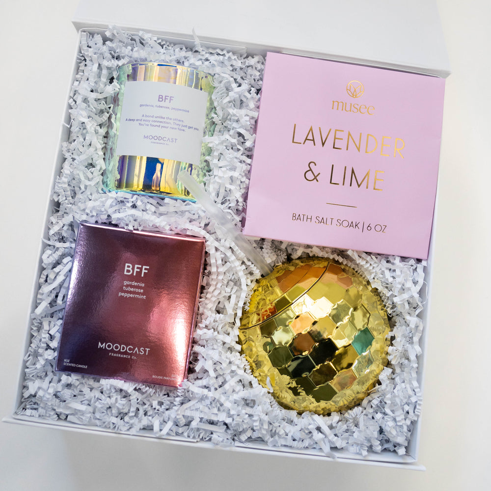 Digital photograph of a curated gift box featuring a pink Musee Lavender & Lime bath soak, an iridescent Moodcast BFF candle jar, and a shiny gold disco ball tumbler nestled in white crinkle paper