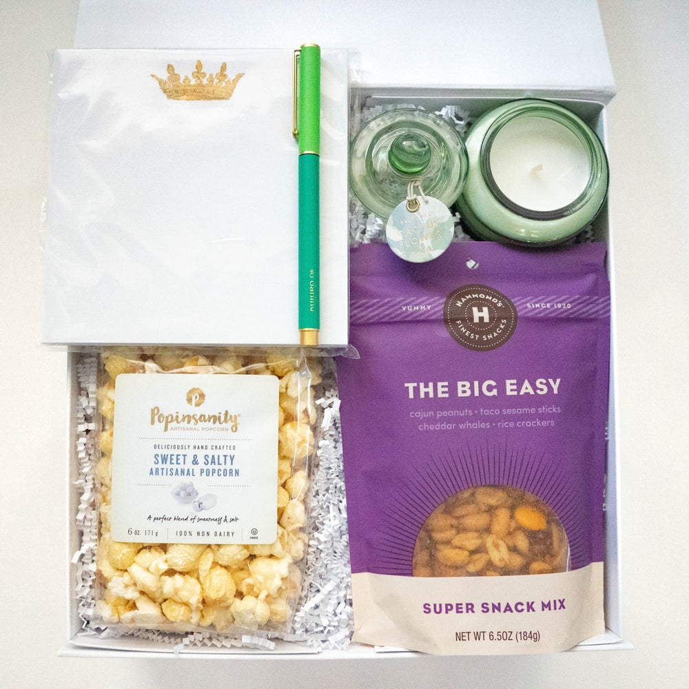 Photograph of a curated gift box containing a vibrant purple bag of Hammond's snacks, a clear bag of Popinsanity popcorn, a bright green pen, and a white envelope with a gold crown logo resting on white crinkle paper