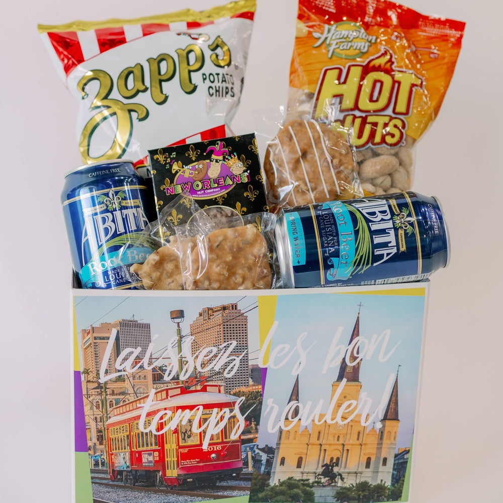 Digital photograph of a New Orleans themed snack box featuring a red streetcar and St. Louis Cathedral landmarks filled with Zapp's potato chips, blue Abita Root Beer cans, and local pralines beneath the phrase Laissez les bon temps rouler