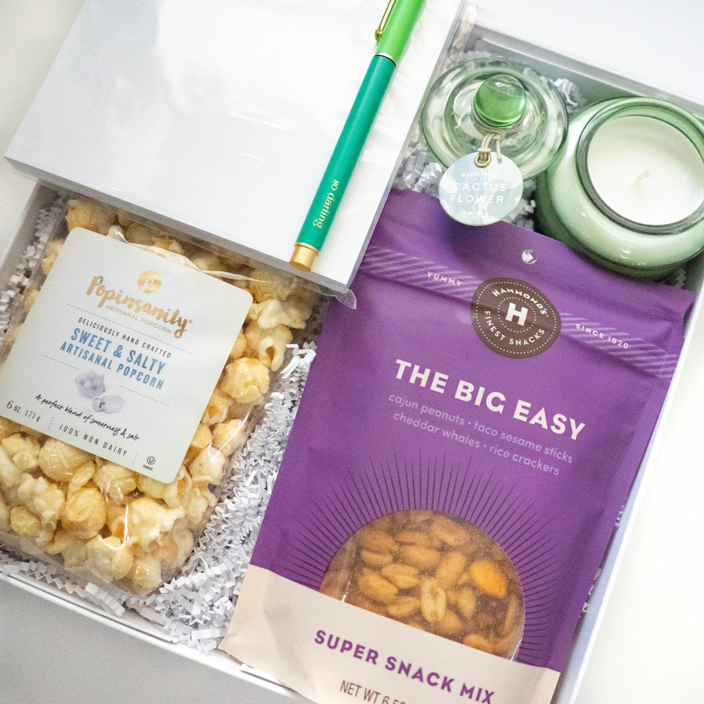 Photograph of a curated gift box featuring a purple bag of Hammond’s snack mix and Popinsanity popcorn nestled next to a light green Cactus Flower candle and a vibrant teal pen on white crinkle paper filler