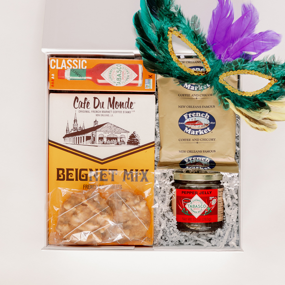 Studio photograph of a New Orleans themed gift box containing Café Du Monde beignet mix featuring the St. Louis Cathedral, a vibrant green and purple feathered Mardi Gras mask with gold sequins, and a classic red bottle of Tabasco sauce