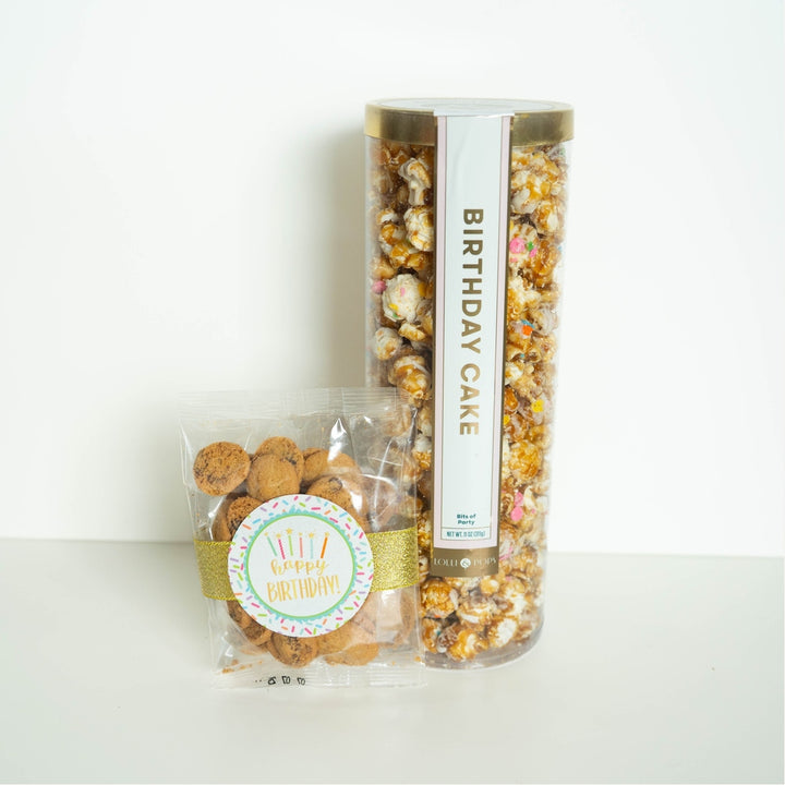 Studio photograph by Lolli &amp; Pops of a tall canister of Birthday Cake popcorn and a clear bag of mini chocolate chip cookies on a white background, showcasing colorful sprinkles, a shimmering gold glitter ribbon, and a round Happy Birthday sticker with pastel candles