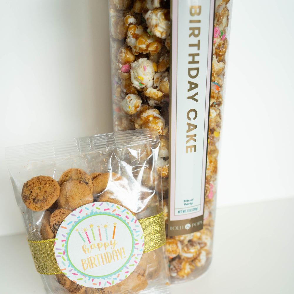 Photograph from Lolli & Pops of a tall cylinder of gourmet Birthday Cake popcorn and a small bag of mini chocolate chip cookies on a white surface, featuring a gold glitter ribbon, a colorful sprinkle-themed Happy Birthday sticker, and popcorn clusters drizzled with white icing and rainbow sprinkles