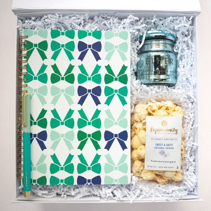 Photograph of a curated gift box featuring a notebook with a repeating teal and navy bow pattern, a matching teal pen with gold accents, an Ether light blue glass candle, and a bag of Popinsanity Sweet &amp; Salty popcorn nestled in white crinkle paper