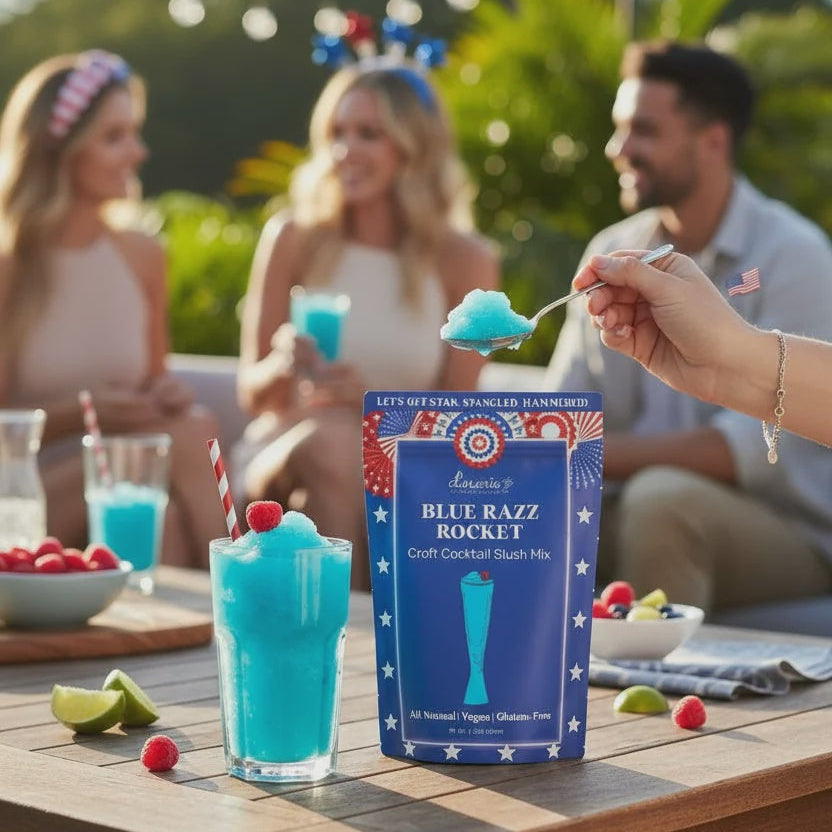 Commercial photograph of Blue Razz Rocket cocktail slush mix on an outdoor wooden table featuring a vibrant blue frozen drink topped with a red raspberry, a spoon lifting a scoop of ice, and patriotic American flag motifs during a festive gathering