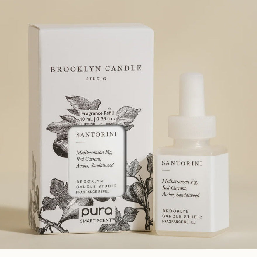 Product photograph by Brooklyn Candle Studio of a Pura Santorini fragrance refill and box featuring black-and-white botanical fig sketches, minimalist typography, and a list of scent notes including Red Currant and Amber against a cream background