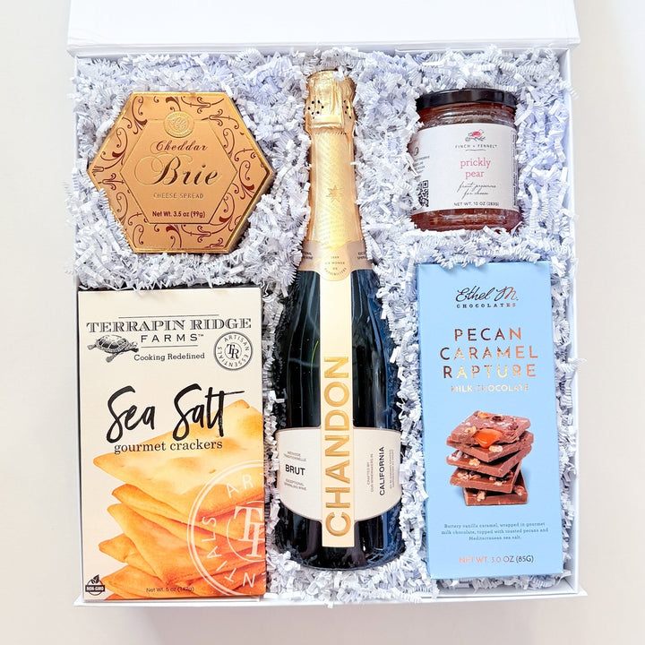 An open gift box filled with white crinkle paper and containing a bottle of Chandon sparkling wine, a box of Terrapin Ridge Farm Sea Salt, a jar of NutraSweet Caramel Sauce, and a round wooden plaque reading &
