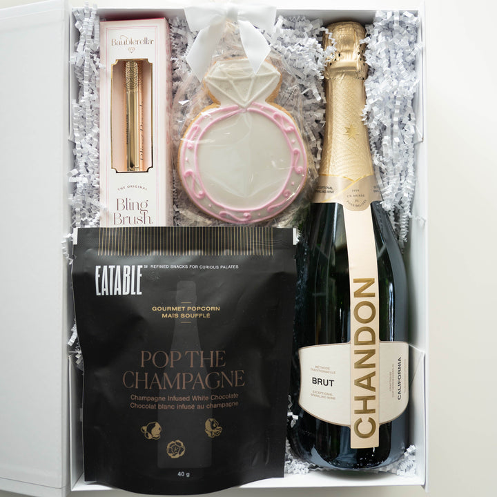 Photograph of a luxury celebration gift box containing a bottle of Chandon Brut sparkling wine, Eatable gourmet popcorn, and a Baublerella Bling Brush nested in white crinkle paper next to a large sugar cookie shaped like an engagement ring with pink icing accents