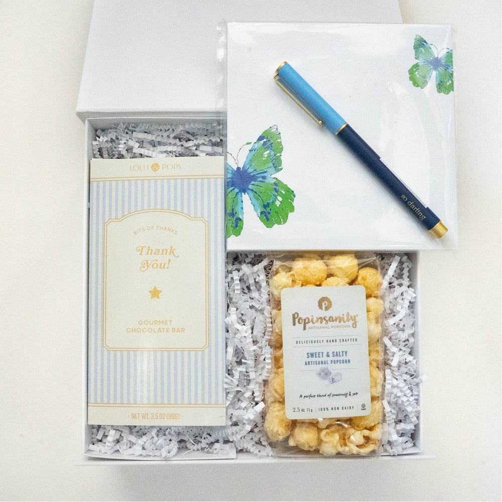 Photograph of a curated gift box featuring a Lolli & Pops gourmet chocolate bar, Popinsanity sweet and salty popcorn, a notepad with vibrant blue and green butterfly illustrations, and a gold-accented blue pen resting on white crinkle paper bedding