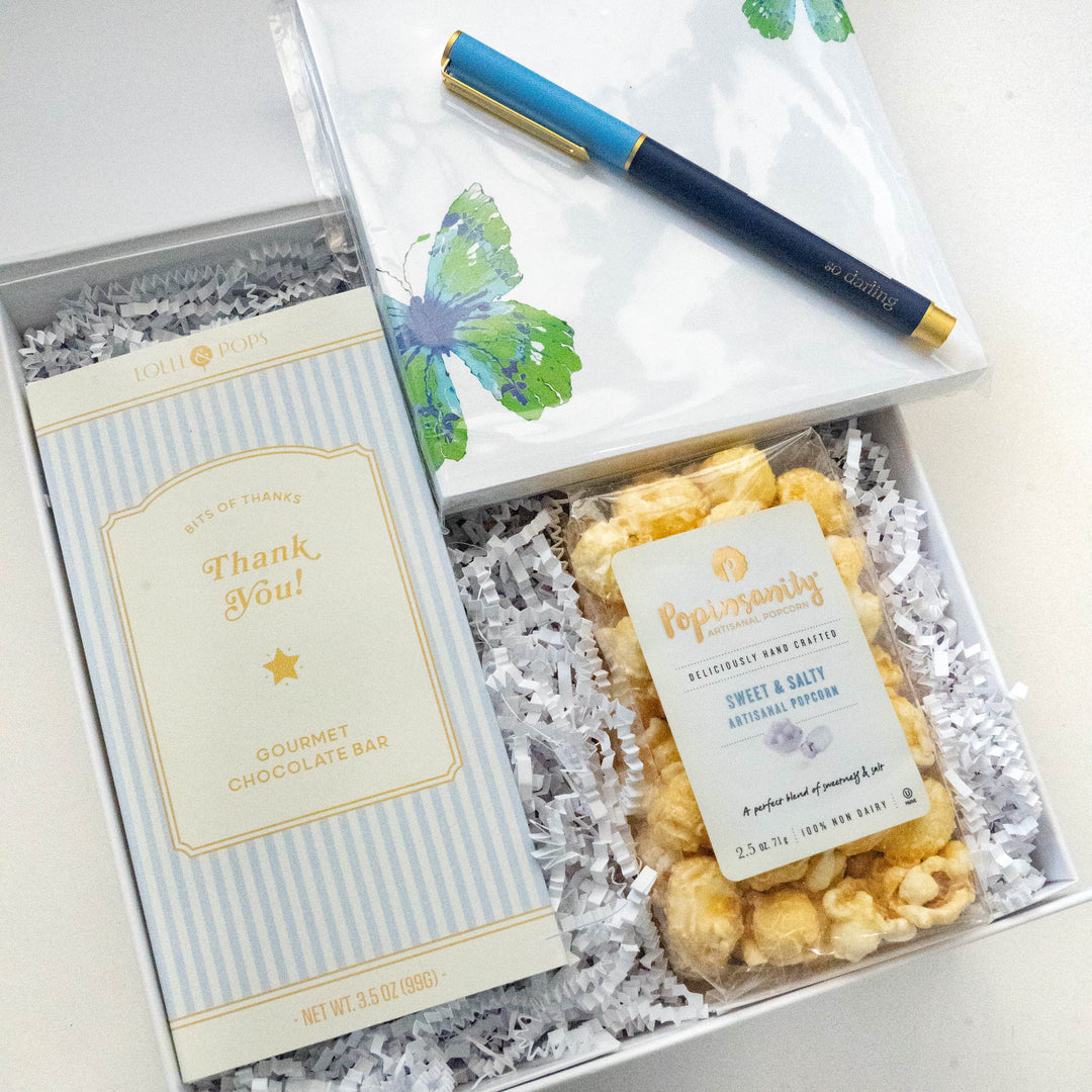 Photograph of a Lolli &amp; Pops and Popinsanity thank you gift set featuring a light blue striped chocolate bar, artisanal sweet and salty popcorn, and a watercolor butterfly notepad with a navy blue so darling pen nestled in white crinkle paper