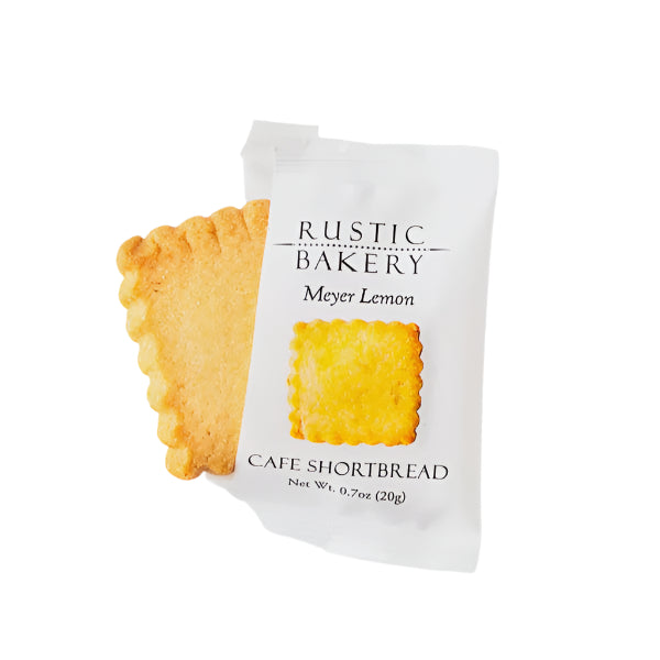 Commercial studio photograph by Rustic Bakery of a Meyer Lemon Cafe Shortbread cookie with scalloped edges emerging from its white matte packaging against a white background, featuring a golden-brown texture and the iconic black Rustic Bakery logo text