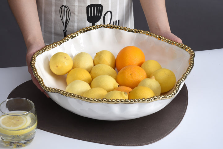 Photograph of a wavy white ceramic fruit bowl with a gold beaded rim filled with lemons and oranges held by a person in a striped apron next to a glass of lemon water