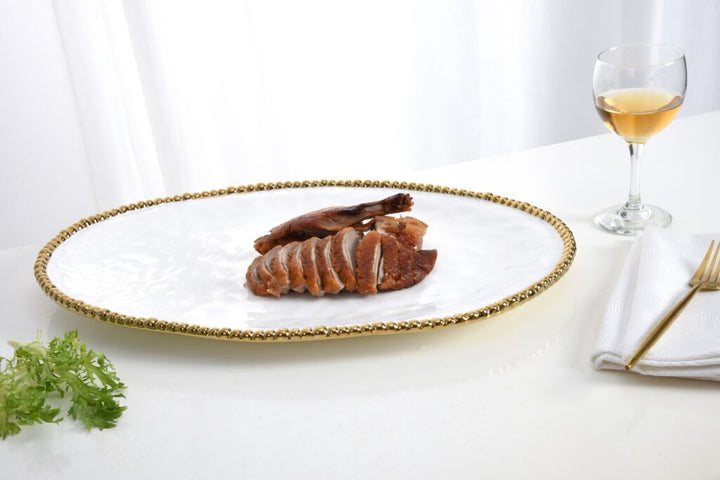 Photograph of sliced roasted duck with crispy skin served on a white textured oval platter featuring a gold beaded rim next to a glass of white wine and a gold fork on a white tabletop
