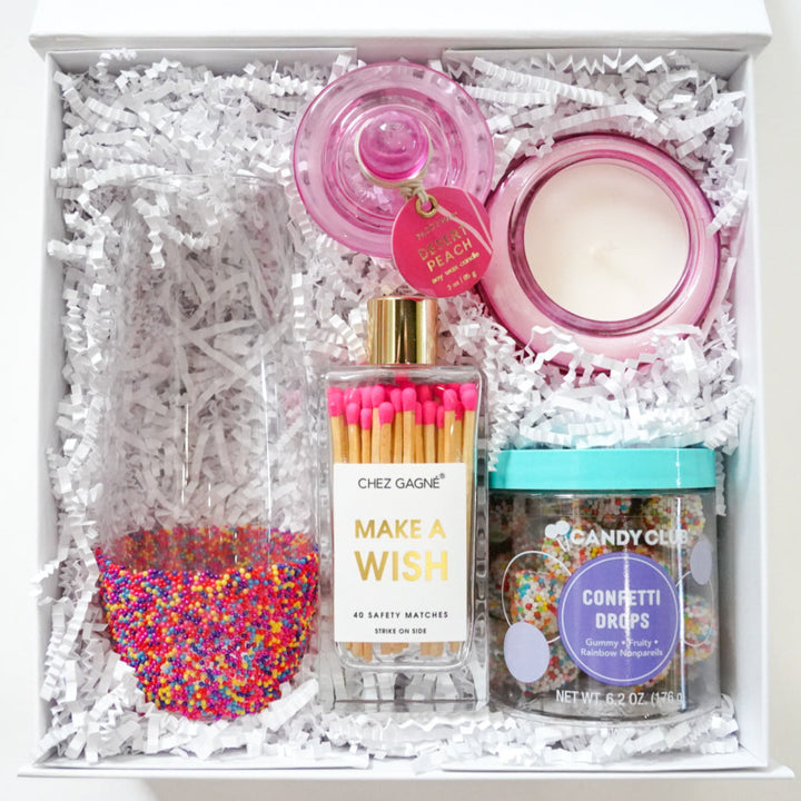Photograph of a curated Chez Gagné celebration gift box featuring a wine glass dipped in multicolored sprinkles, a pink Desert Peach jar candle, and a bottle of bright pink-tipped matches nestled on white shredded paper