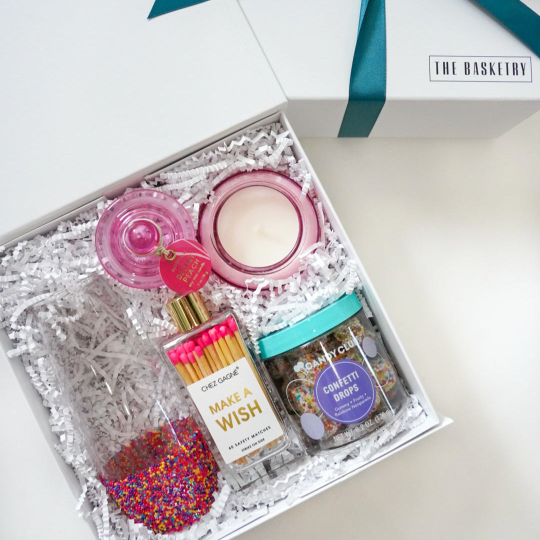 Photograph by The Basketry of a curated white gift box containing a pink glass candle, pink-tipped Chez Gagné matches, and a jar of Candy Club confetti drops nestled in white crinkle paper with vibrant multi-colored nonpareils