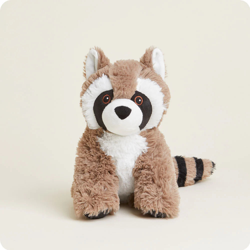 A plush raccoon toy sitting in a studio against a neutral cream background featuring a signature black eye mask with orange-rimmed eyes, a white fluffy chest, and a thick striped tail