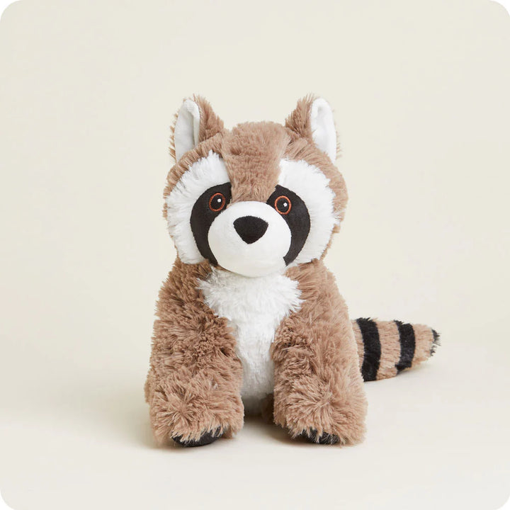 A plush raccoon toy sitting in a studio against a neutral cream background featuring a signature black eye mask with orange-rimmed eyes, a white fluffy chest, and a thick striped tail