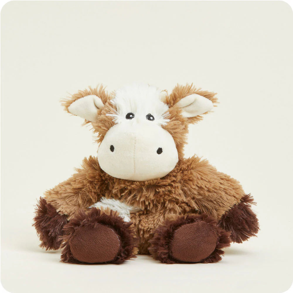 Plush toy of a shaggy brown and white cow sitting against a plain off-white background, featuring a cream-colored face with small black eyes and dark brown hooves