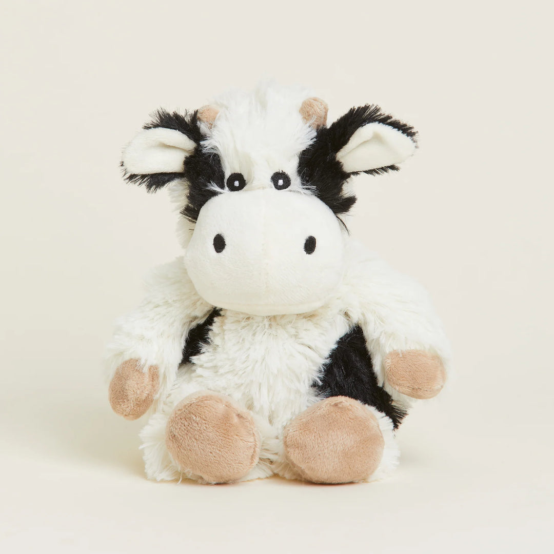 Studio photograph of a Warmies plush cow toy sitting centered on a cream background with fuzzy black and white patches, small tan horns, and soft beige hooves