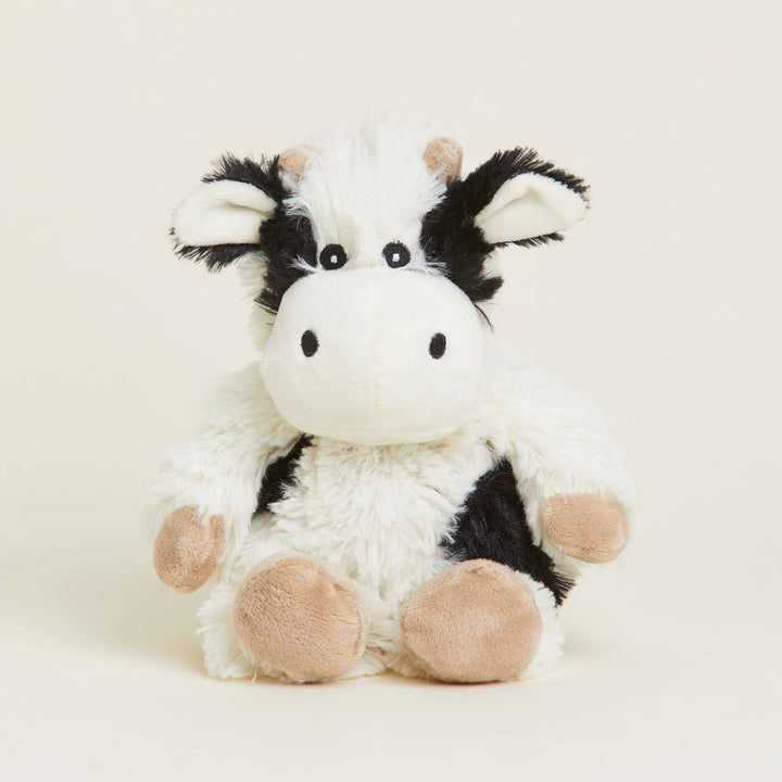 Studio photograph of a Warmies plush cow toy sitting centered on a cream background with fuzzy black and white patches, small tan horns, and soft beige hooves