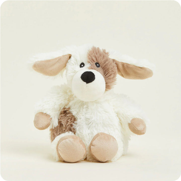 Plush toy of a white and brown spotted puppy dog sitting on a plain cream background featuring floppy ears with tan interiors, a fuzzy brown patch over one eye, and smooth beige paw pads