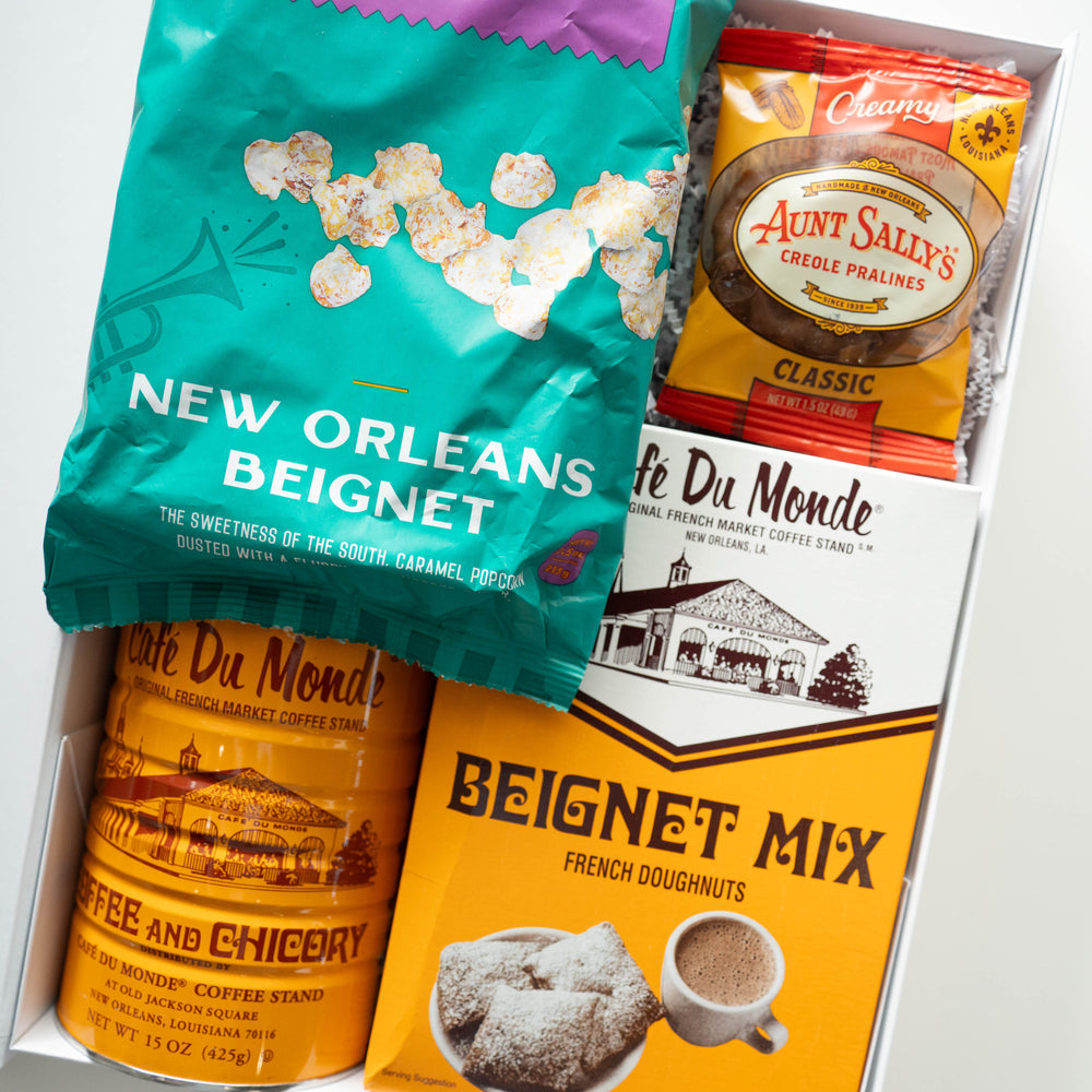 Photograph of a New Orleans snack gift box featuring the iconic Café Du Monde French Market stand illustration on yellow packaging, a vibrant teal bag of beignet popcorn with a trumpet graphic, and a package of Aunt Sally’s Creole Pralines