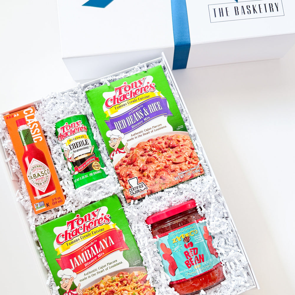 Photograph by The Basketry of a Louisiana-themed culinary gift box containing iconic Tabasco sauce and Tony Chachere’s Creole mixes nestled in white crinkle paper with vibrant green packaging and a royal blue ribbon