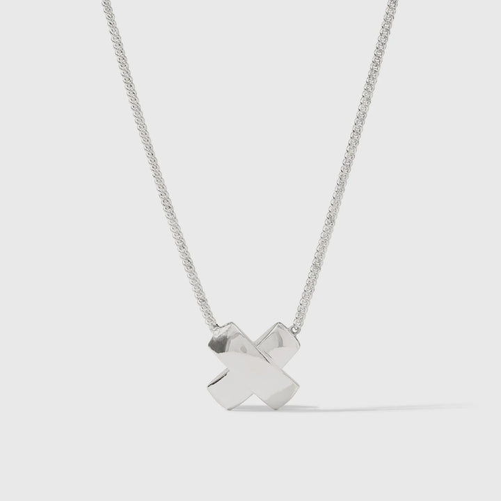 Sterling silver necklace by Paloma Picasso featuring a polished X-shaped kiss pendant on a delicate curb chain in a neutral studio setting with reflective highlights and a soft shadow