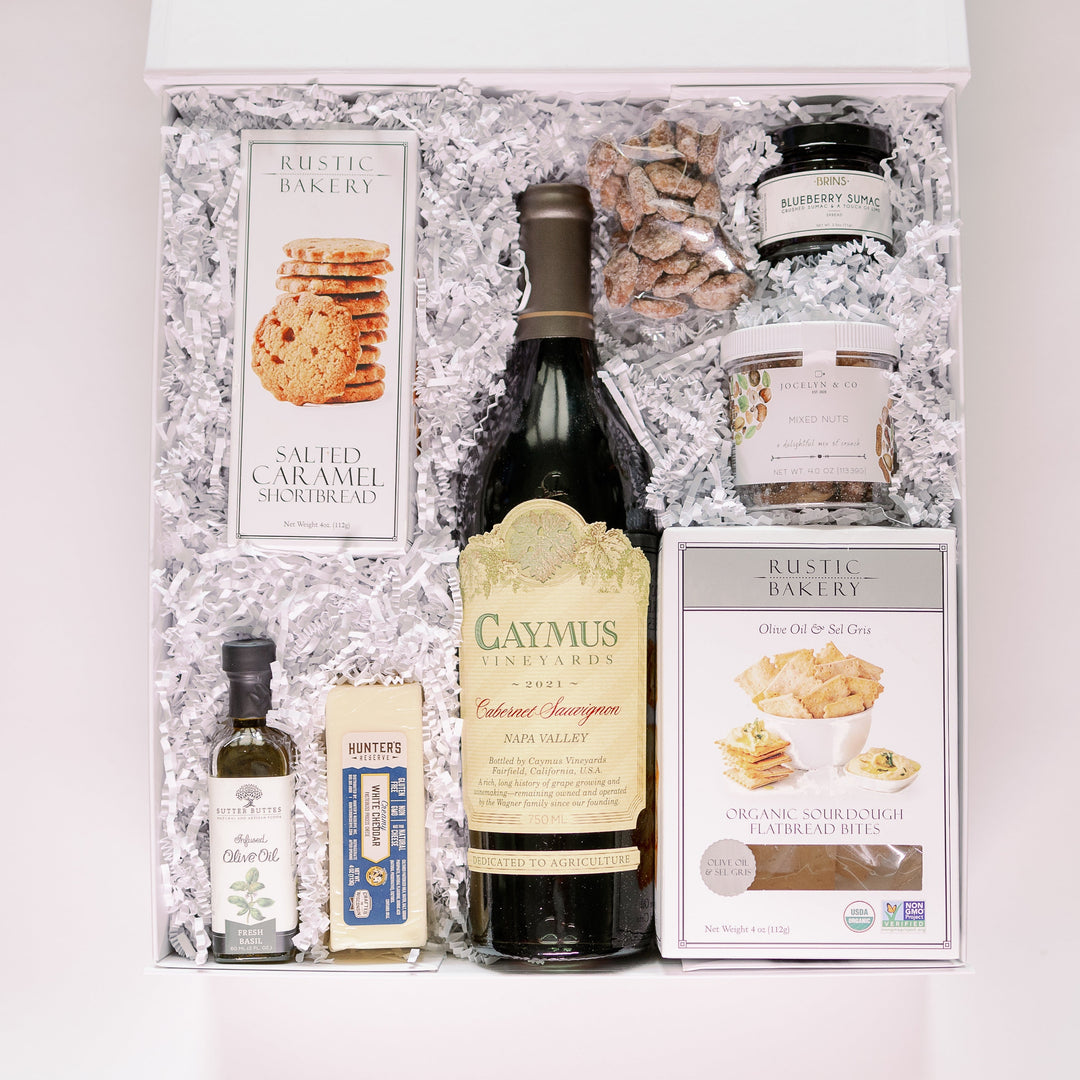 Studio photograph of a luxury gourmet gift box featuring Caymus Cabernet Sauvignon, Rustic Bakery shortbread, and a small jar of blueberry sumac spread nestled in white shredded paper