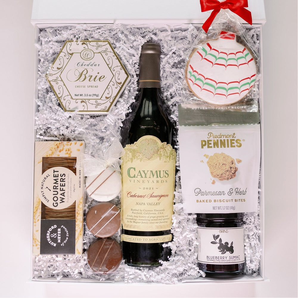 Photograph of a luxury holiday wine gift box featuring a bottle of Caymus Vineyards Cabernet Sauvignon and Piedmont Pennies savory bites arranged on white crinkle paper with a festive red-and-green-iced ornament cookie and a jar of blueberry sumac jam