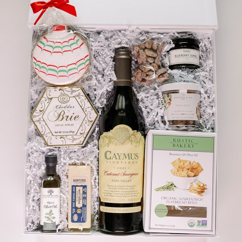 Photograph of a gourmet wine and food gift box featuring a central bottle of Caymus Vineyards Cabernet Sauvignon alongside Rustic Bakery sourdough flatbread, a colorful red and green ornament-shaped sugar cookie, and artisanal cheese nestled in white crinkle-cut paper filler with a vibrant red bow