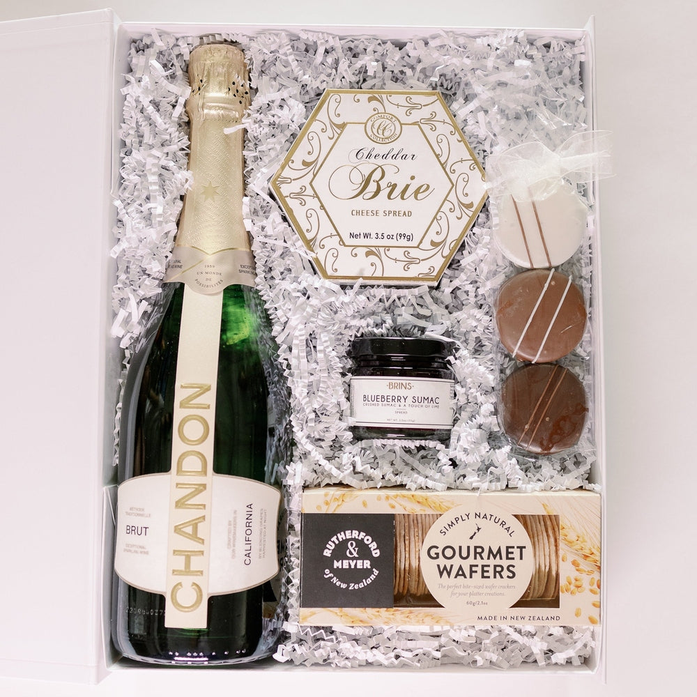 Digital photograph of a luxury Chandon champagne gift box featuring a gold-foiled Brut bottle, Rutherford & Meyer gourmet wafers, a hexagonal brie spread, and three striped chocolate-covered treats nestled in white shredded paper inside a white gift box
