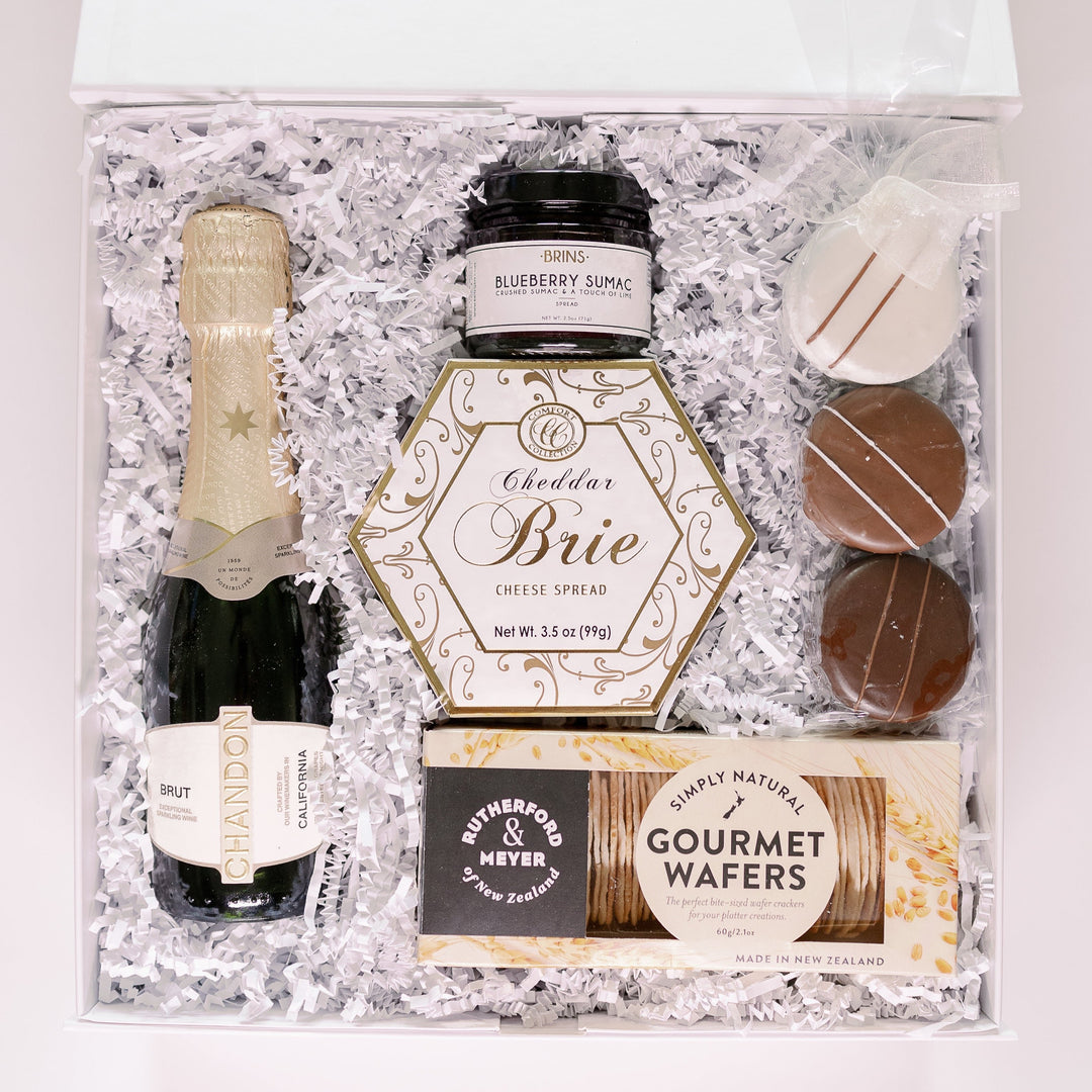 Product photograph of a luxury gourmet gift box arranged in white shredded paper featuring a bottle of Chandon Brut, hexagonal-shaped Comfort Collection Cheddar Brie, and Rutherford & Meyer wafers accented by a jar of blueberry sumac and chocolate-dipped cookies with decorative white stripes